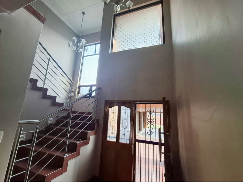 Spacious double-volume entrance hall featuring a timber staircase with stainless steel balustrades, security gate, and natural light from high-level windows.