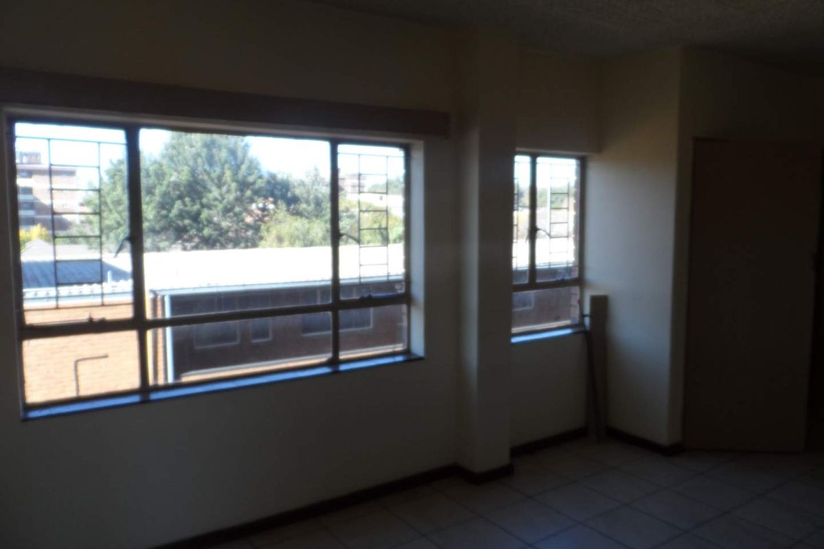 An empty living area featuring large windows with security bars, tiled flooring, and neutral wall finishes, offering natural light and a view of the surroundings.