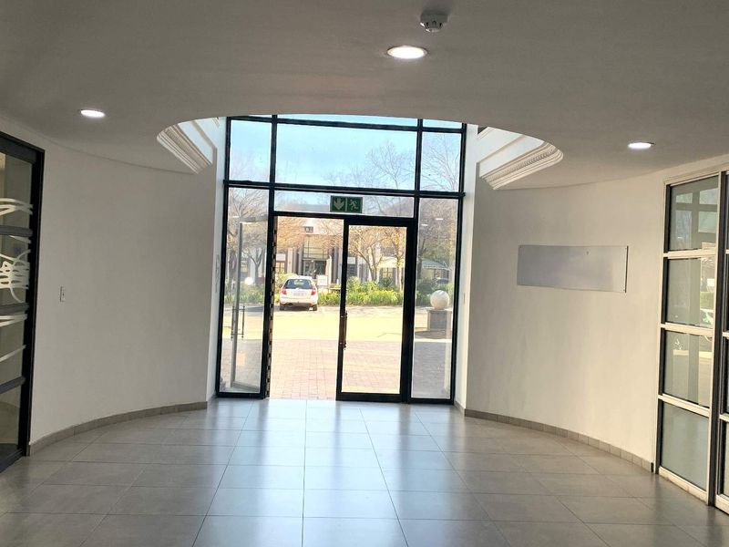 A bright and modern office entrance lobby featuring tiled flooring, recessed ceiling lights, and large glass doors providing natural light and exterior access.