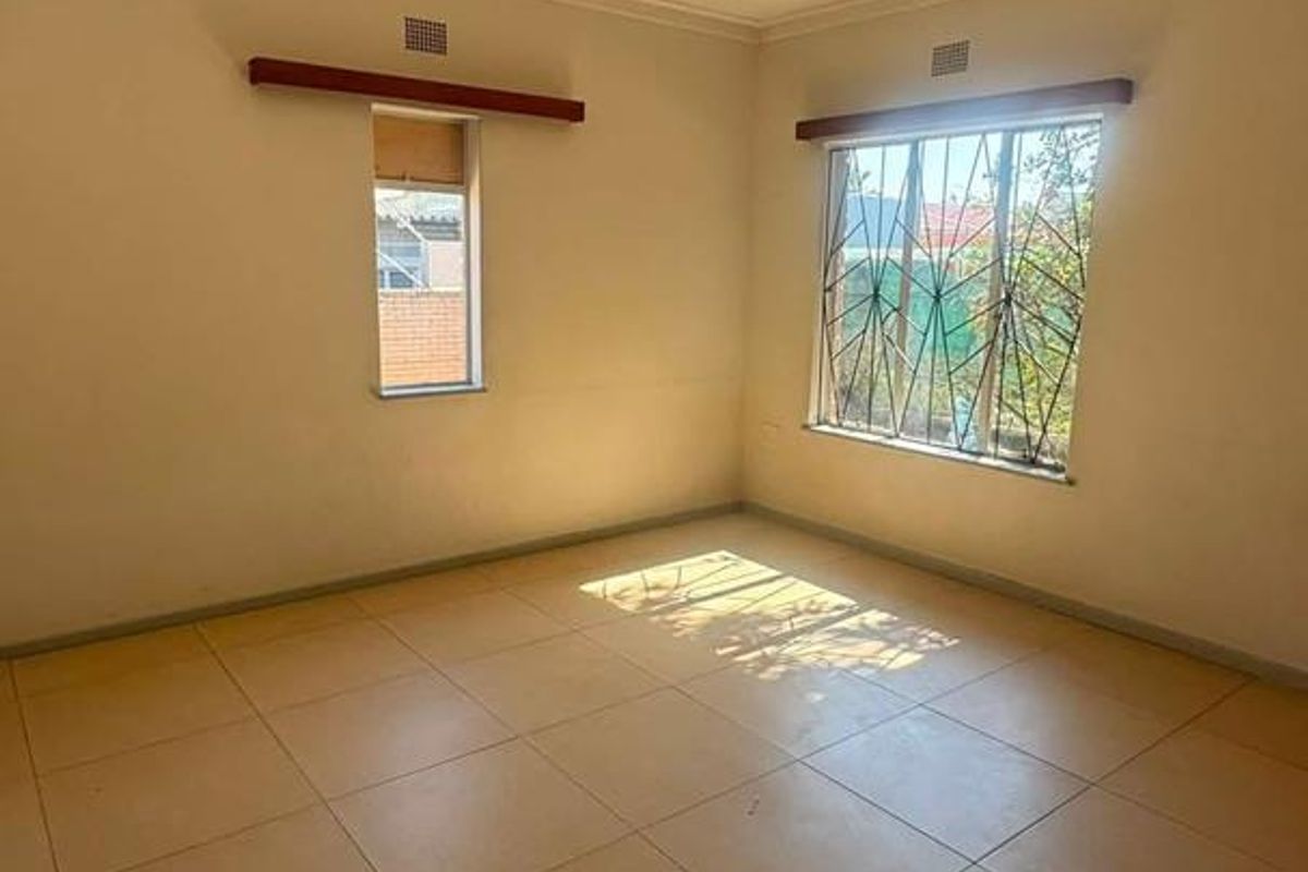 A bright, empty bedroom featuring neutral tiled flooring, security bars on the windows, and simple curtain rails.
