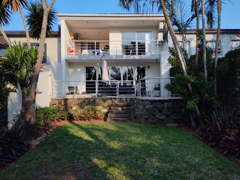 Rear view of the property featuring a landscaped garden, stone retaining wall, ground-level patio, and an upper-level balcony with metal railings.