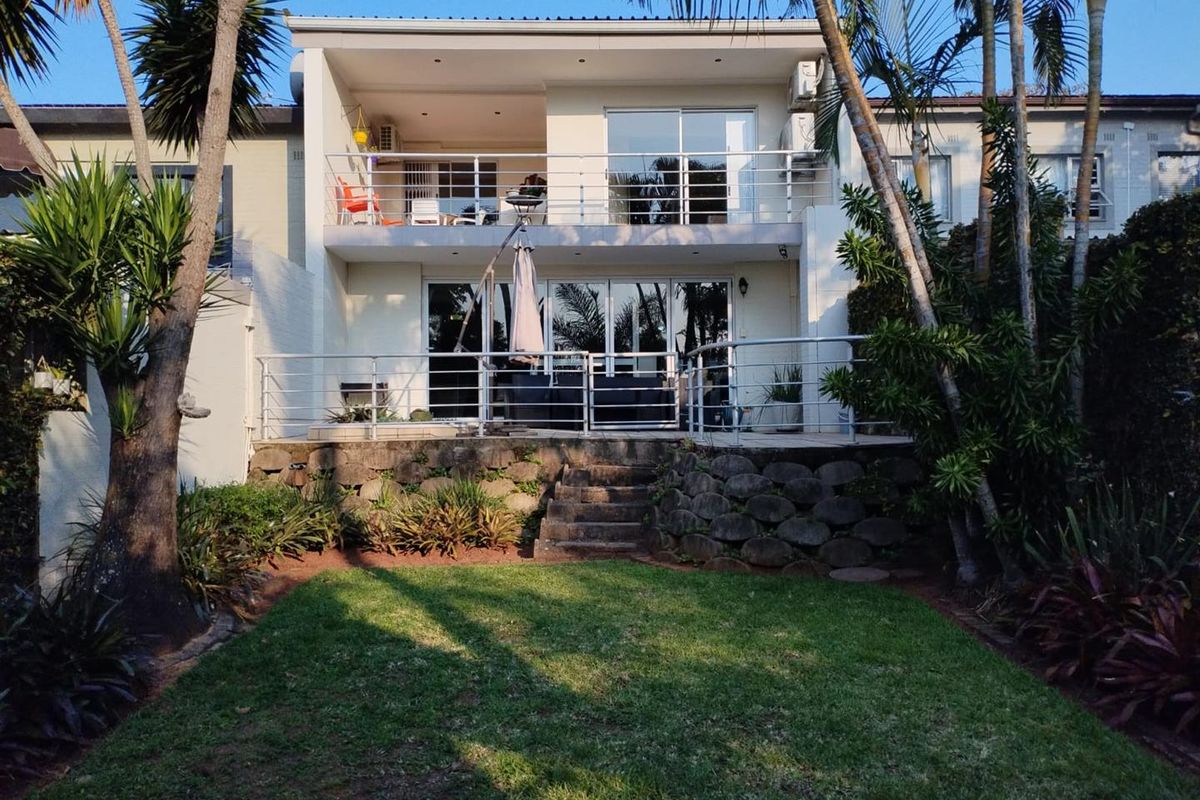 Rear view of the property featuring a landscaped garden, stone retaining wall, ground-level patio, and an upper-level balcony with metal railings.
