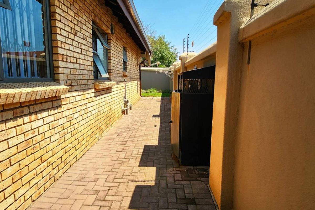 A paved side walkway along the exterior of the townhouse, featuring face-brick walling, security bars on windows, and a perimeter boundary wall.