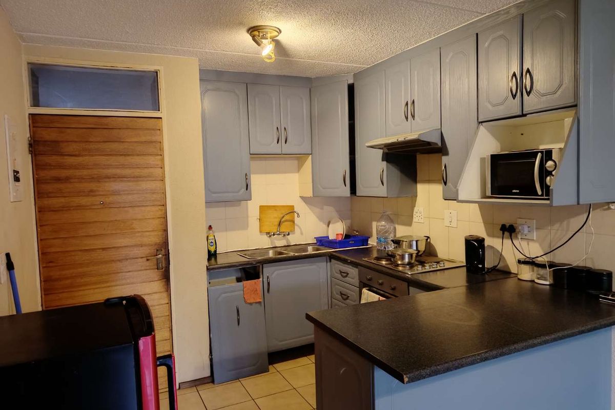 Compact kitchen featuring grey painted cabinetry, laminate countertops, built-in oven, hob, and extractor fan, with tiled flooring and direct access to the entrance.