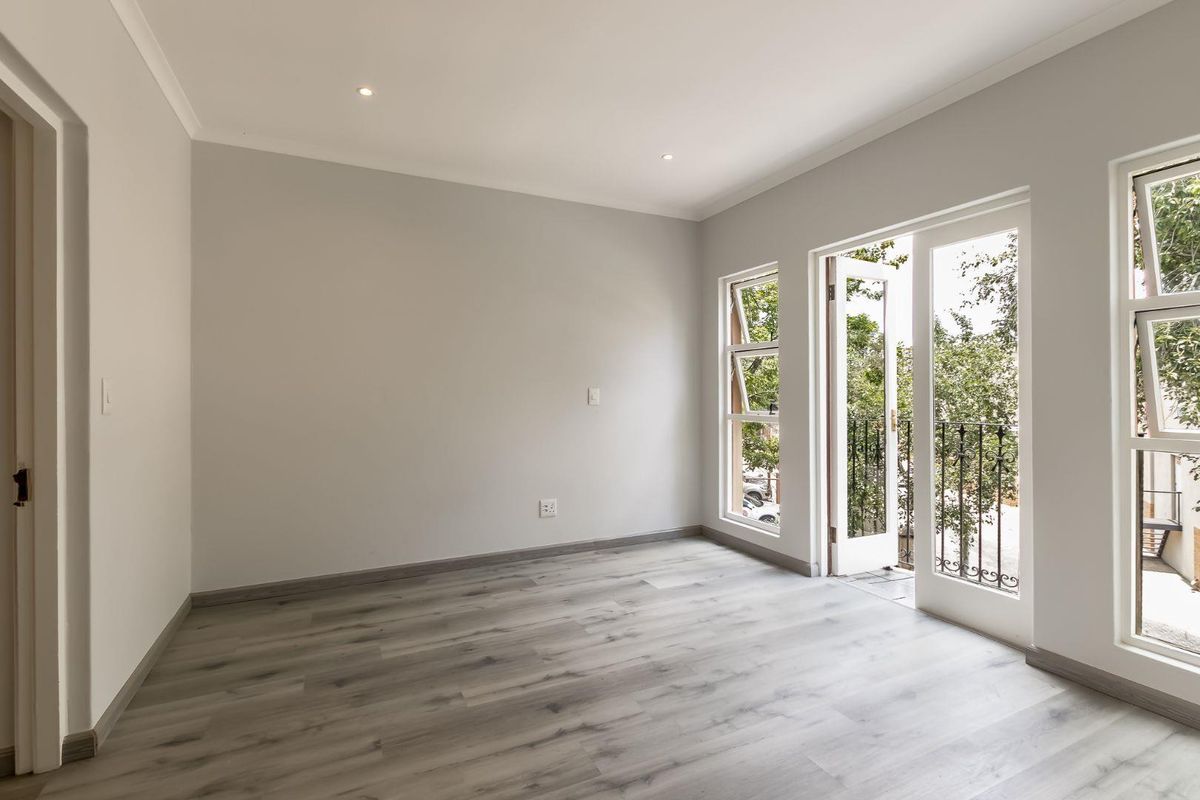 A bright, empty bedroom featuring modern wood-look laminate flooring, recessed ceiling lighting, and a glass door opening onto a small balcony.