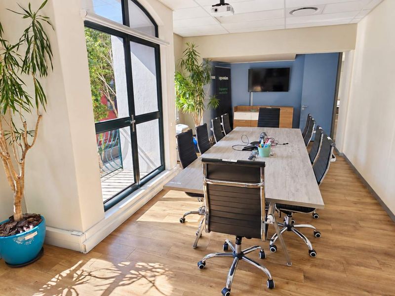 A well-lit office boardroom featuring laminate flooring, a suspended ceiling, large windows, and integrated audiovisual equipment including a wall-mounted television and projector.