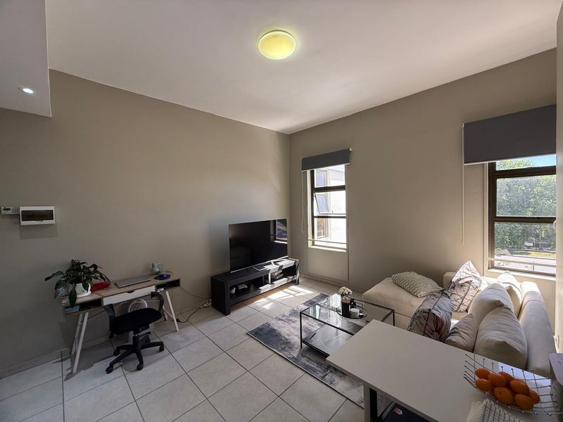 A well-lit living room featuring neutral wall tones, tiled flooring, and large windows with roller blinds, offering a comfortable and functional living space.