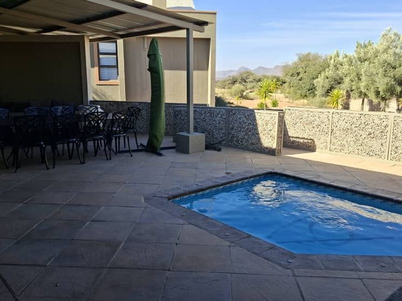 Outdoor entertainment area featuring a covered patio, paved flooring, and a swimming pool, enclosed by a stone-clad boundary wall with scenic views.