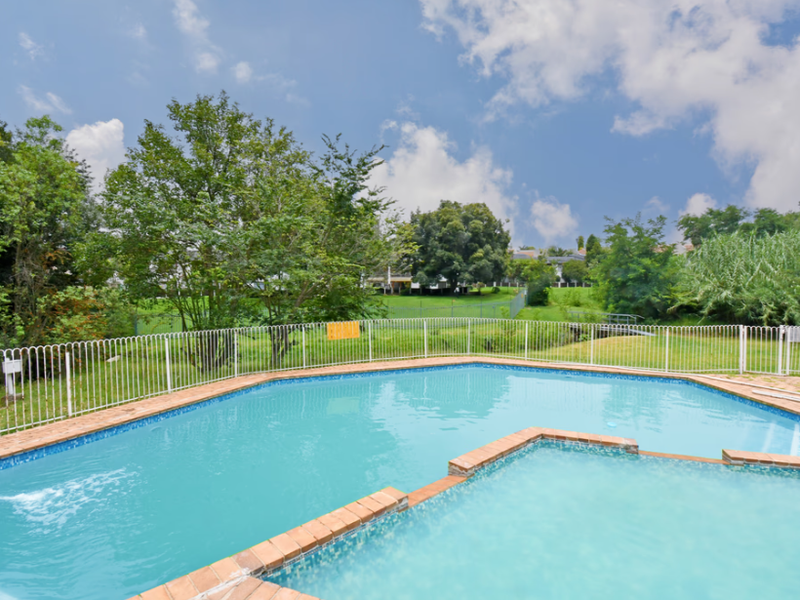 A well-maintained outdoor swimming pool area featuring brick coping and secure metal perimeter fencing, set within a landscaped garden environment.