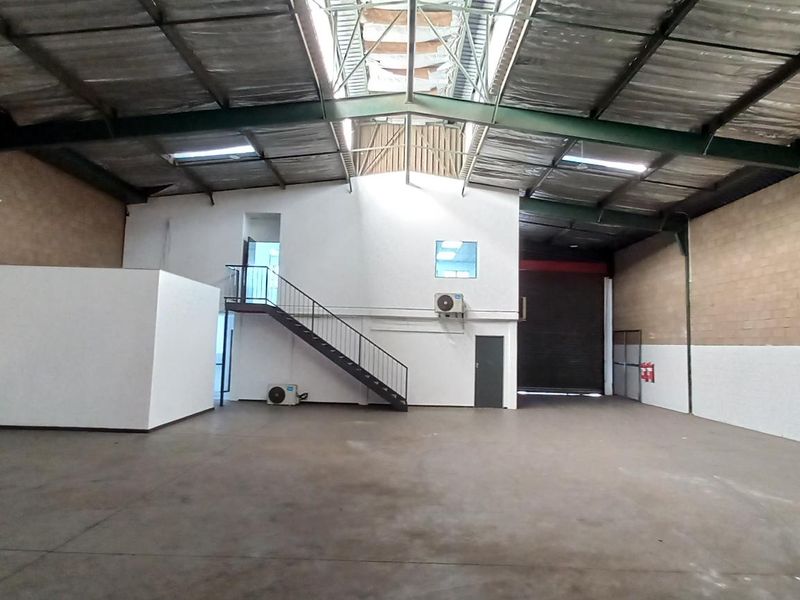 Spacious industrial warehouse featuring high ceilings, exposed steel trusses, a mezzanine level with staircase, roller shutter door, and concrete flooring.