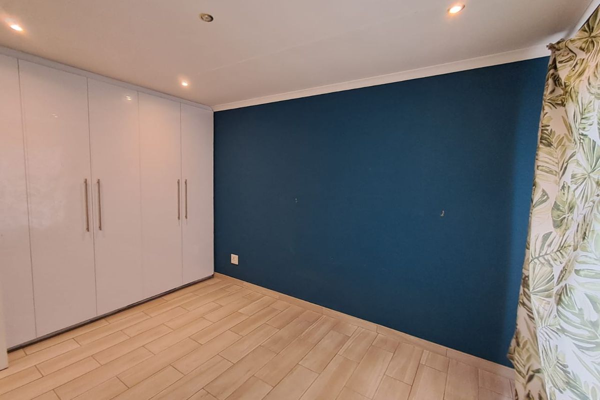 A bright bedroom featuring modern wood-look tile flooring, recessed ceiling lighting, and a large wall-to-wall built-in wardrobe with a sleek white finish.