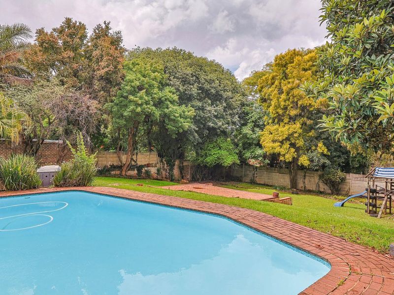 A private backyard featuring a swimming pool with brick coping, surrounded by a lush lawn and mature trees with a perimeter boundary wall.