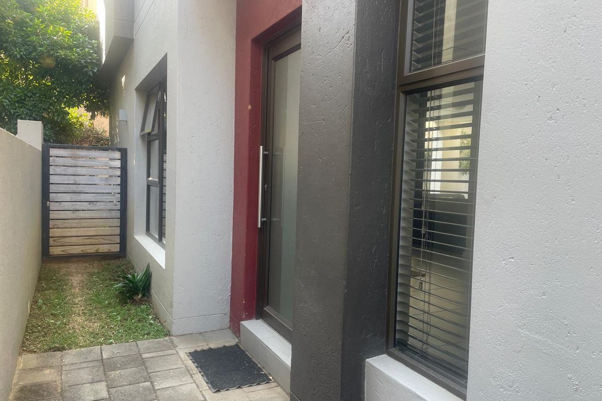 A side entrance area featuring paved flooring, a modern glass door, and a small garden section with a wooden gate.