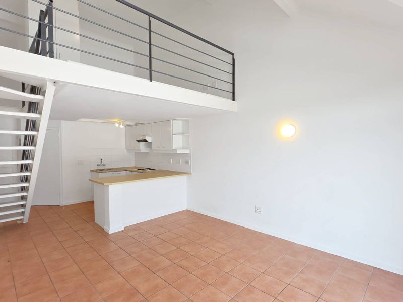 Open-plan living area featuring a mezzanine level, metal staircase, and a compact kitchen with built-in cabinetry and tiled flooring throughout.