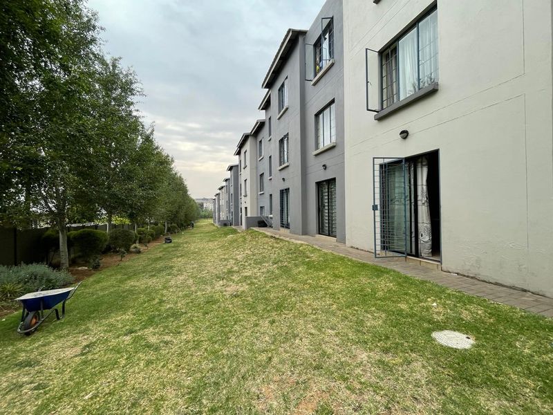 Rear view of the apartment complex showing the ground floor units with security gates, paved walkways, and a shared lawn area.