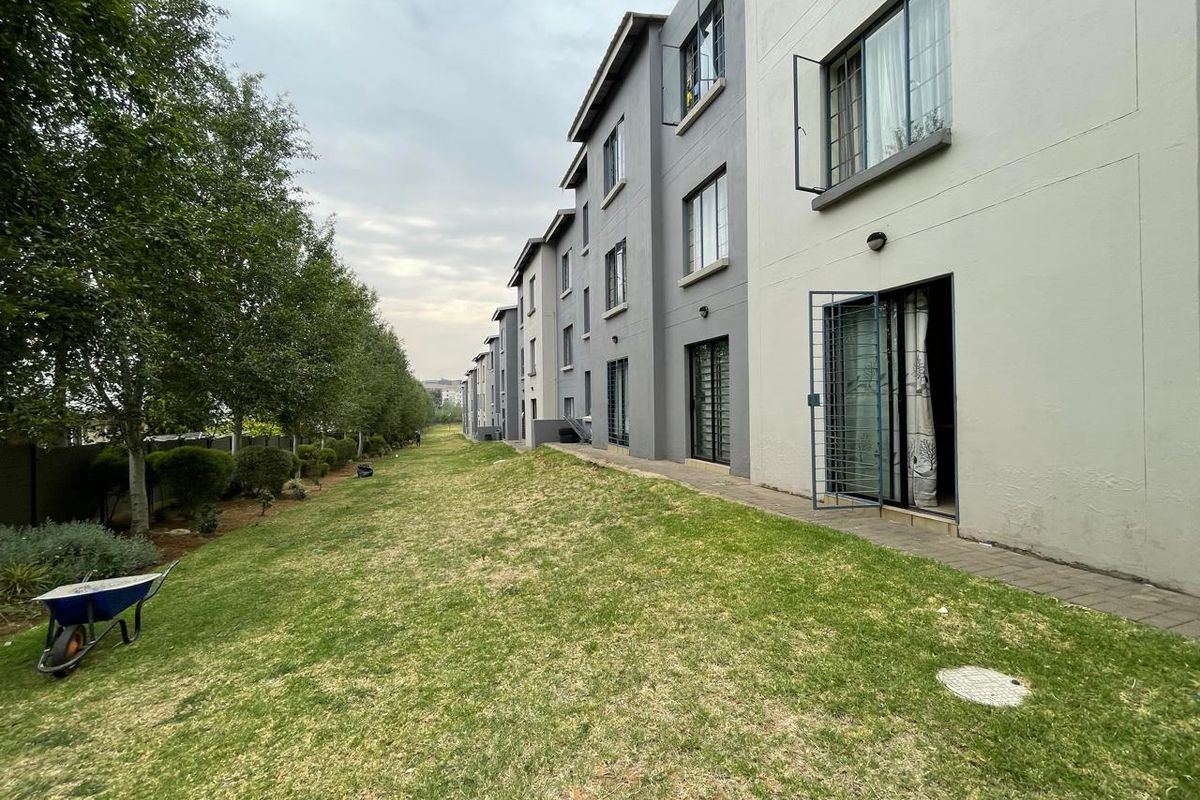 Rear view of the apartment complex showing the ground floor units with security gates, paved walkways, and a shared lawn area.