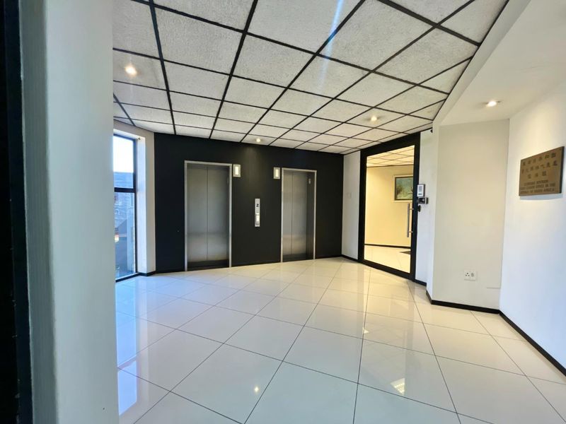 A modern office building lobby featuring a double elevator bank, polished tile flooring, and a suspended ceiling with recessed lighting.