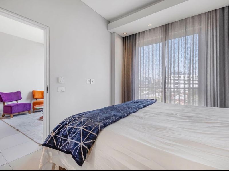 A bright bedroom featuring tiled flooring, recessed ceiling lighting, and large windows with curtain tracks, offering a view into an adjacent living space.