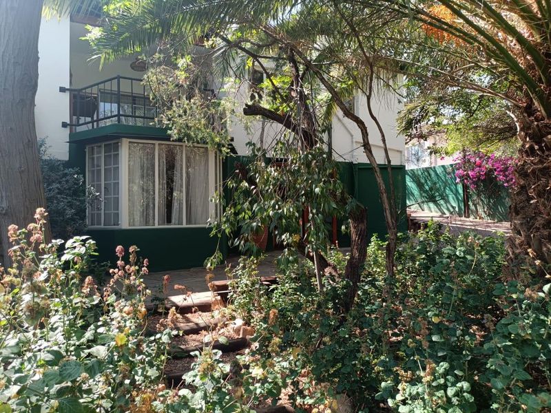 Rear view of the apartment featuring a paved patio area, a ground-floor bay window, and an upper-level balcony with metal railings, surrounded by established garden greenery.