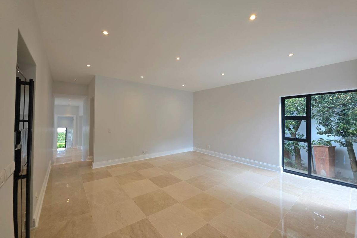A bright, spacious, and empty living area featuring polished stone tile flooring, recessed downlights, and a large window overlooking a private garden.