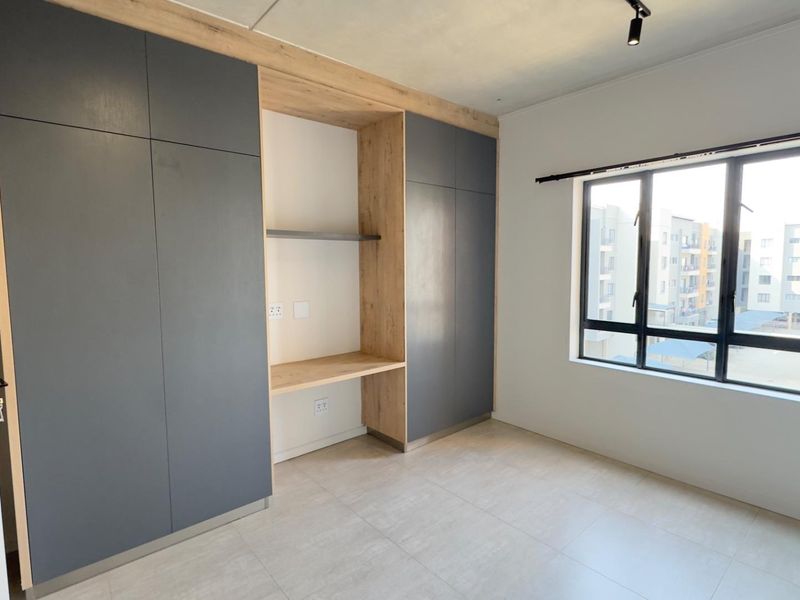 A modern, empty bedroom featuring sleek grey built-in cupboards with an integrated desk space, tiled flooring, and large aluminium-framed windows.