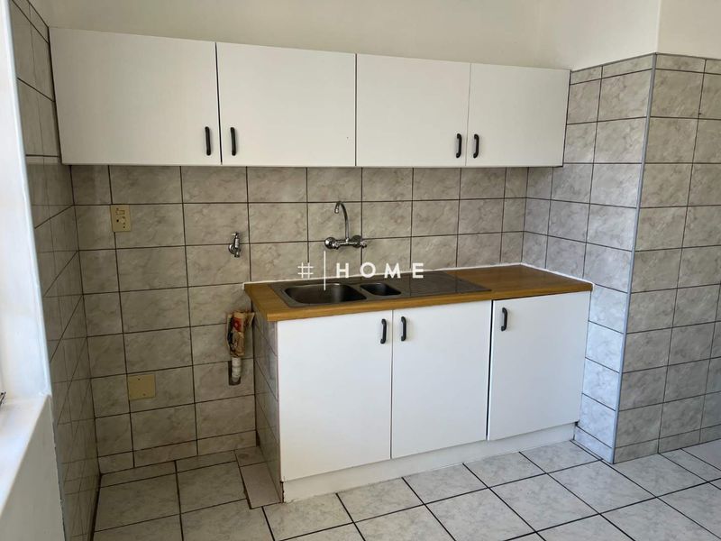 A compact kitchen area featuring white built-in cabinetry, a laminate countertop with a stainless steel sink, and tiled wall and floor finishes.