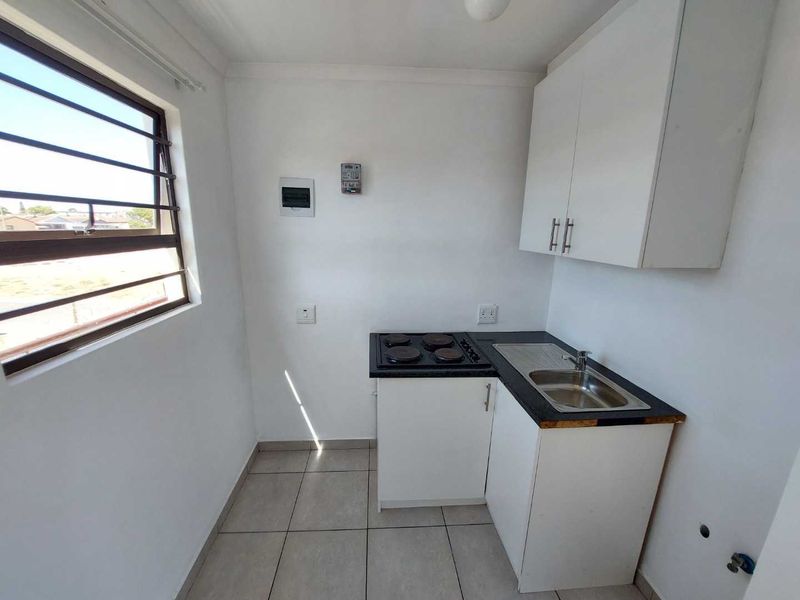 Compact kitchen featuring white cabinetry, a stainless steel sink, and an electric hob, with tiled flooring and a window with security bars.
