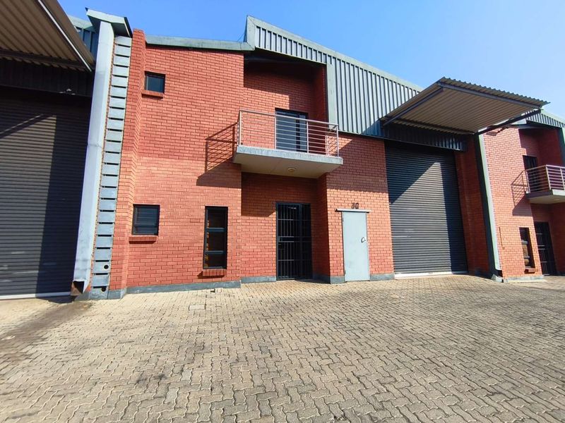 Front elevation of an industrial unit featuring red brick construction, roller shutter doors, and a small balcony, set within a paved industrial complex.