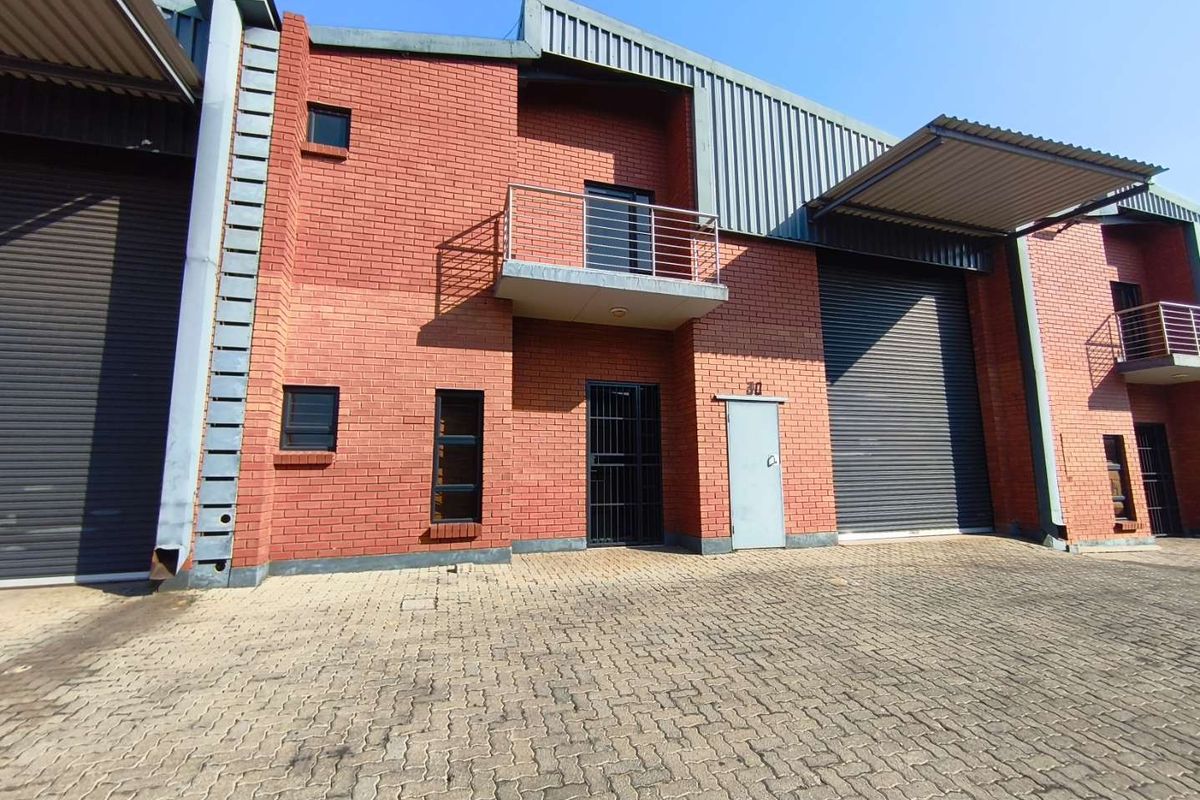Front elevation of an industrial unit featuring red brick construction, roller shutter doors, and a small balcony, set within a paved industrial complex.