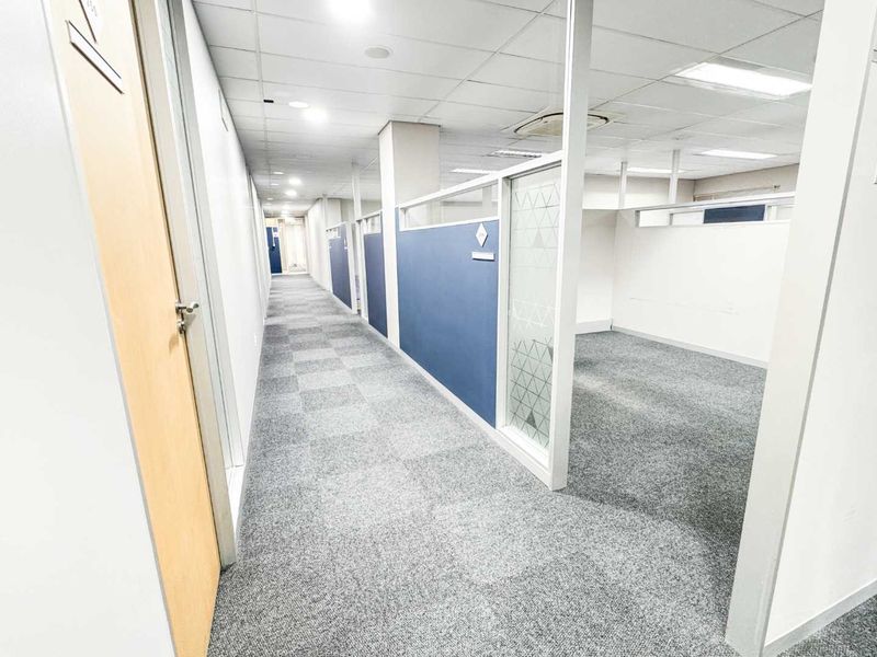 A bright, modern office corridor featuring glass partitions, carpeted flooring, and a suspended ceiling with integrated lighting, providing a professional and clean workspace environment.