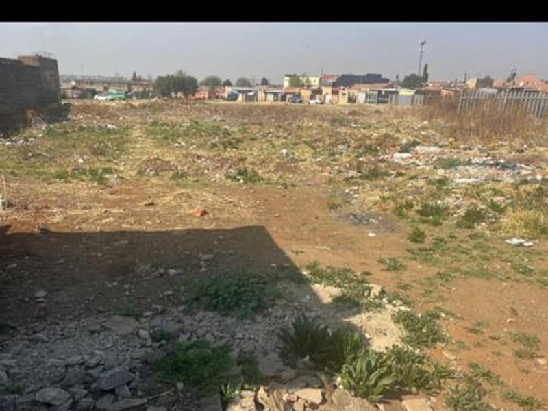 A view of a vacant residential plot featuring uneven terrain, scattered debris, and partial boundary walling in a suburban setting.