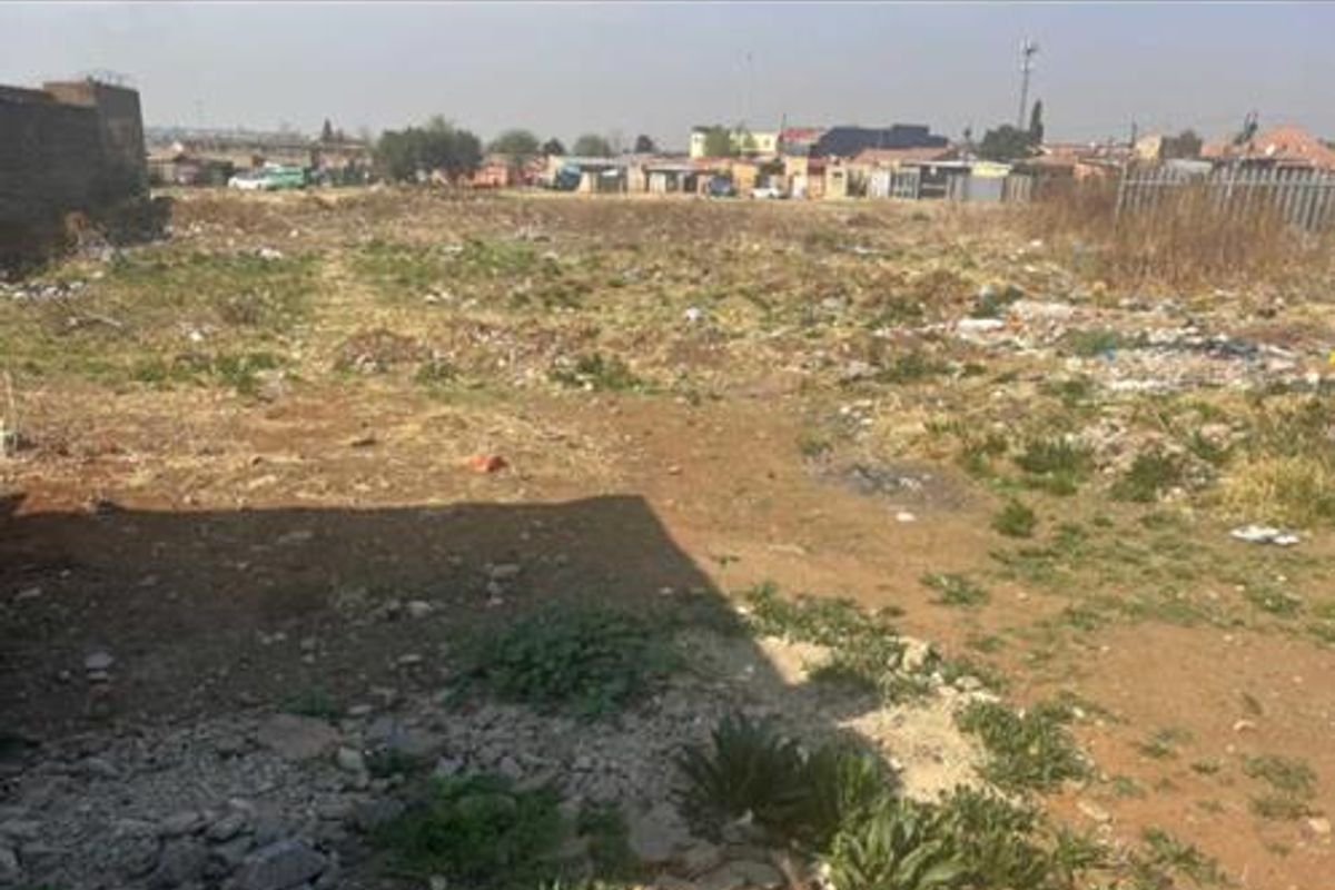A view of a vacant residential plot featuring uneven terrain, scattered debris, and partial boundary walling in a suburban setting.