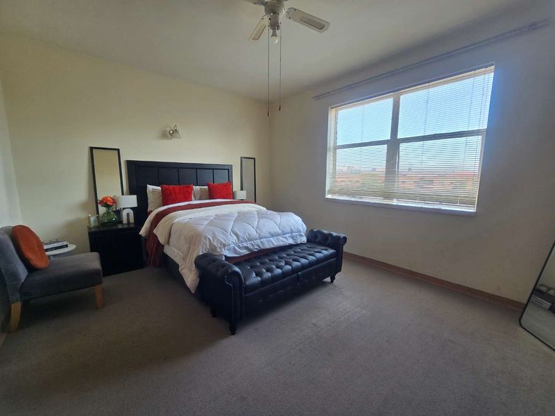 A spacious bedroom featuring neutral walls, carpeted flooring, a ceiling fan, and a large window with venetian blinds providing natural light.