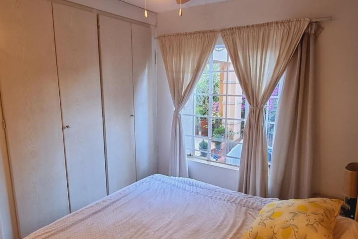 A bedroom featuring built-in cupboards, a ceiling fan, and a window with security bars overlooking the exterior area.