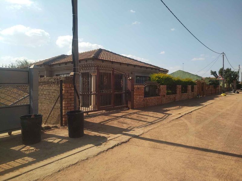 Front exterior view of a single-story house featuring a brick boundary wall, wrought iron gate, and tiled roof, situated on an unpaved street.