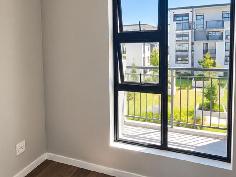 A bright, empty bedroom featuring modern laminate flooring, neutral walls, and a large window with views of the surrounding apartment complex.