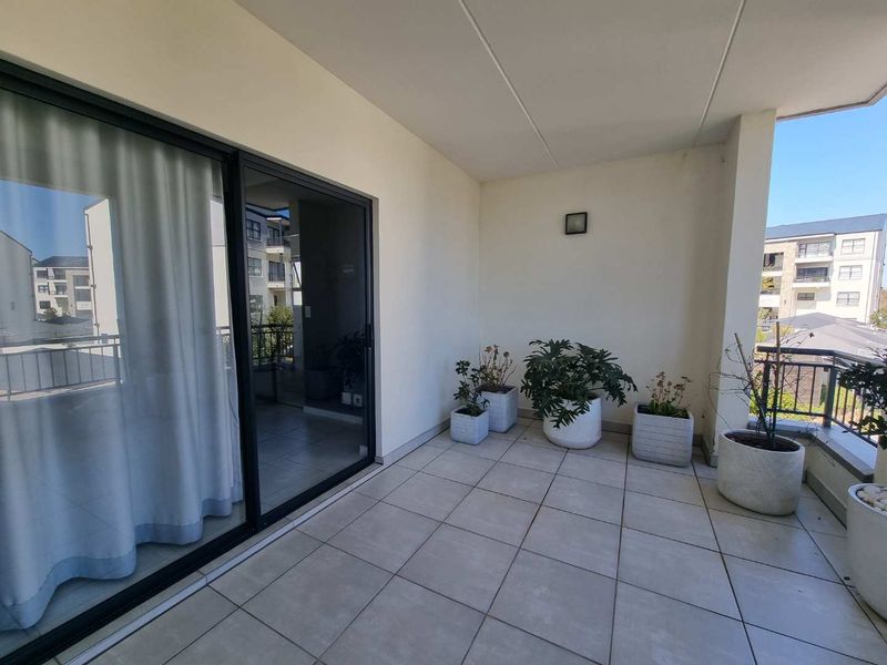 A spacious covered balcony featuring tiled flooring, a modern sliding glass door, and a metal railing, offering a clean and functional outdoor living space.