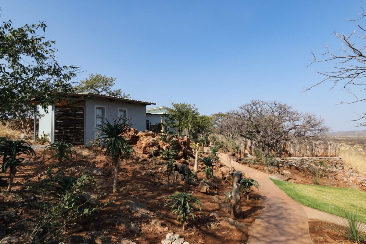 Exterior view of a lodge unit featuring natural rock landscaping, a paved pathway, and indigenous vegetation set against a scenic bushveld backdrop.