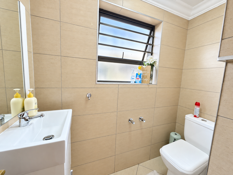 A clean, modern bathroom featuring neutral wall tiles, a wall-mounted basin with chrome fixtures, and a louvered window for natural ventilation.