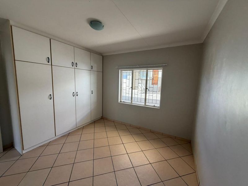 A clean, empty bedroom featuring neutral tiled flooring, built-in storage cupboards, and a window fitted with security bars.