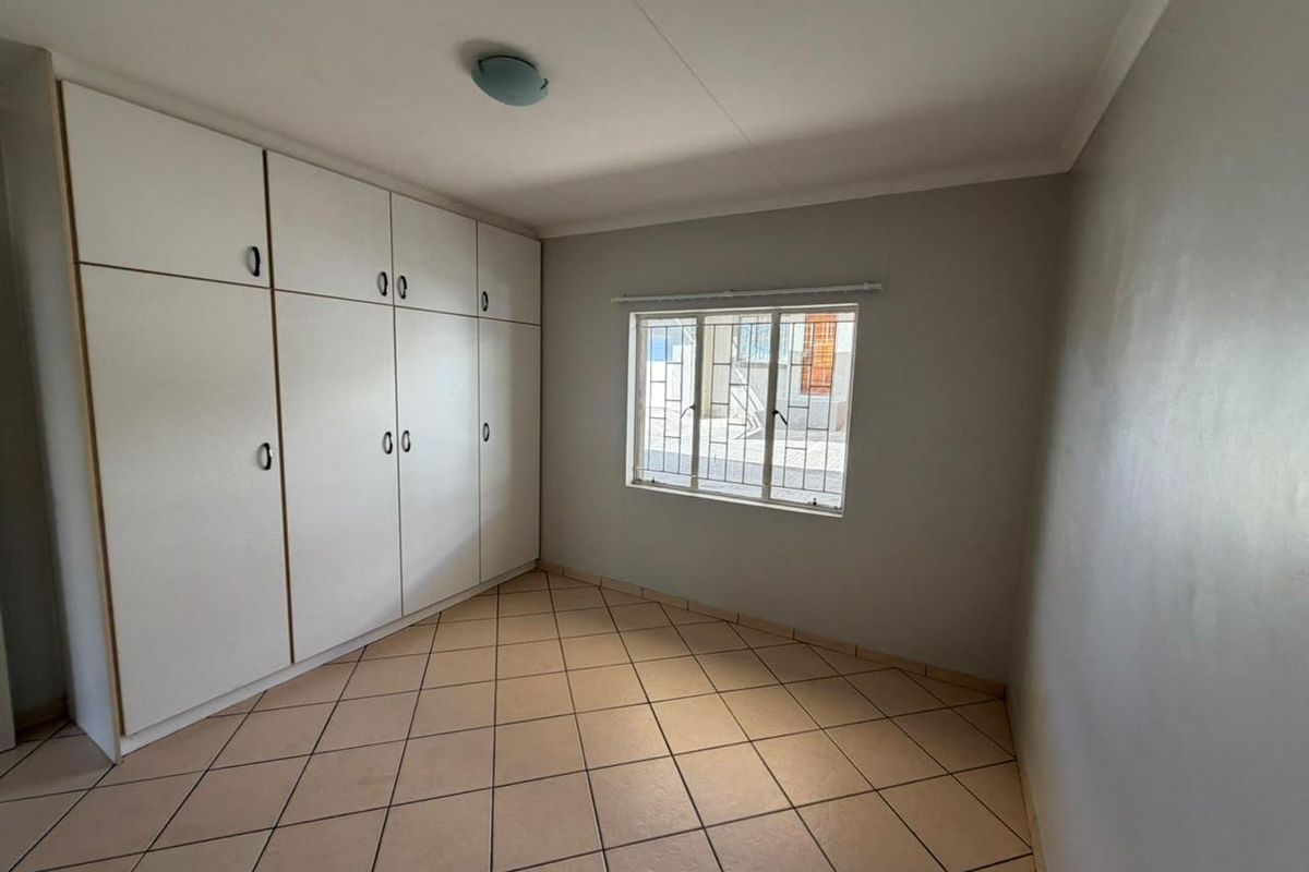 A clean, empty bedroom featuring neutral tiled flooring, built-in storage cupboards, and a window fitted with security bars.