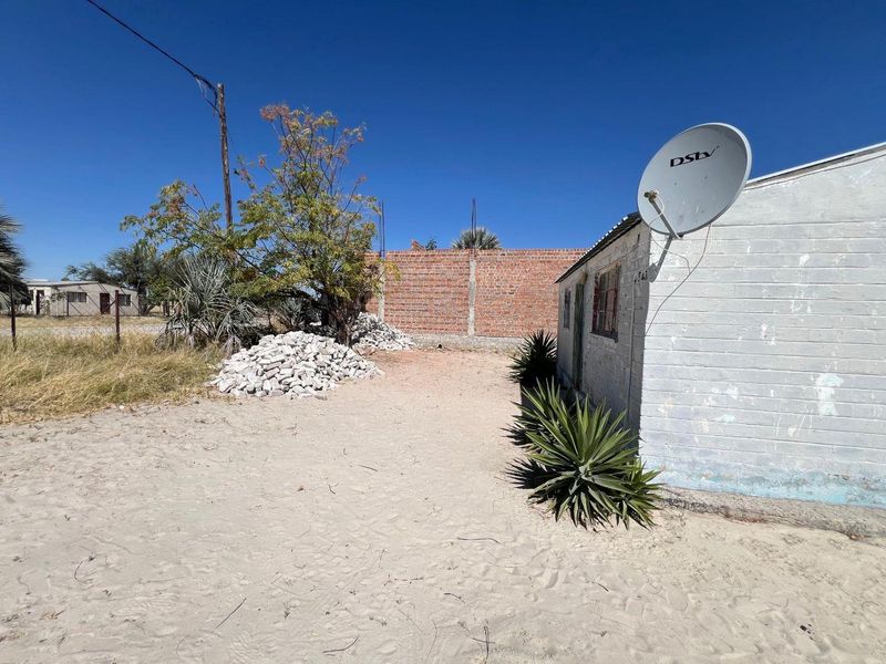 Rear exterior view showing a sandy yard, an unfinished brick boundary wall, and the side elevation of the house with a satellite dish.