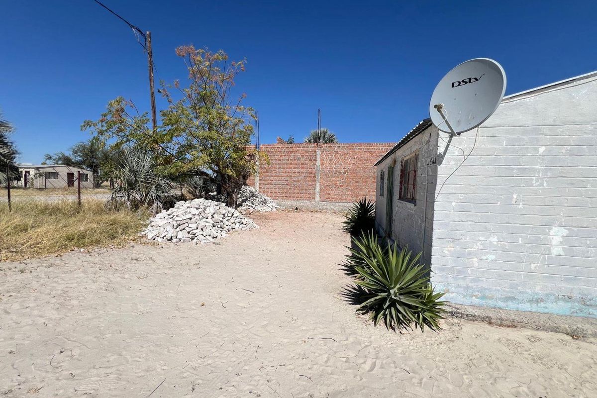 Rear exterior view showing a sandy yard, an unfinished brick boundary wall, and the side elevation of the house with a satellite dish.