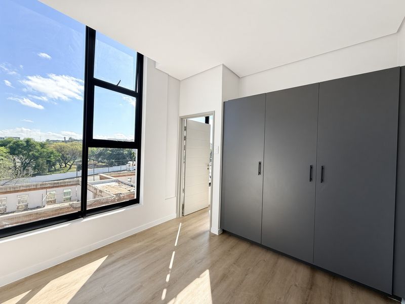 A bright, modern bedroom featuring sleek dark grey built-in cupboards, light laminate flooring, and a large window offering expansive views.