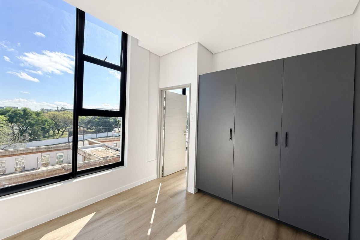 A bright, modern bedroom featuring sleek dark grey built-in cupboards, light laminate flooring, and a large window offering expansive views.