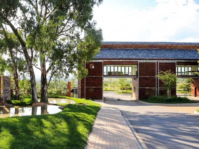The entrance to Blue Hills Equestrian Estate featuring a modern timber-clad gatehouse, paved driveway, landscaped gardens, and a decorative water feature.