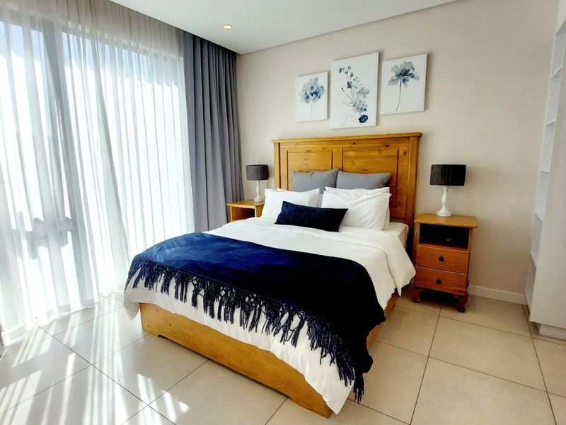 A bright and well-maintained bedroom featuring neutral tiled flooring, recessed ceiling lighting, and a built-in shelving unit with large windows and floor-to-ceiling curtains.