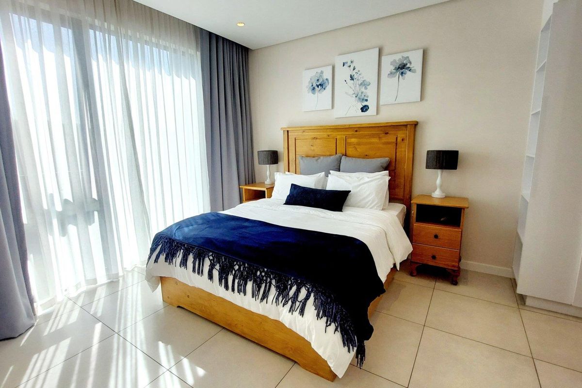 A bright and well-maintained bedroom featuring neutral tiled flooring, recessed ceiling lighting, and a built-in shelving unit with large windows and floor-to-ceiling curtains.