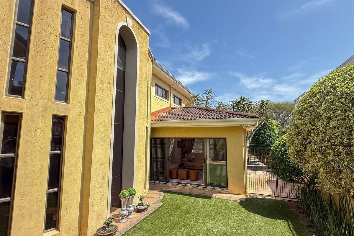 Rear view of the property featuring a covered patio area, yellow stucco facade, tall vertical windows, and a small landscaped garden with artificial turf.