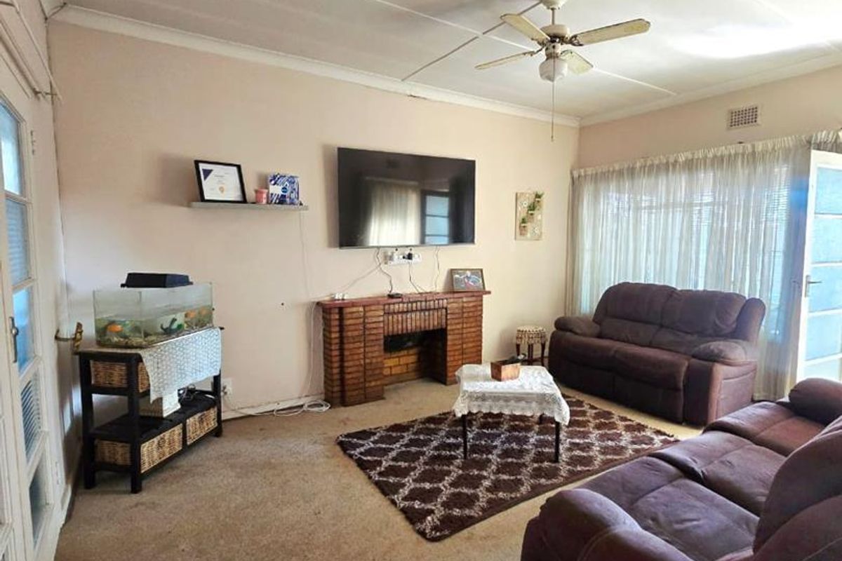 A living room featuring a brick fireplace, ceiling fan, and tiled ceiling, with worn carpeting throughout the space.